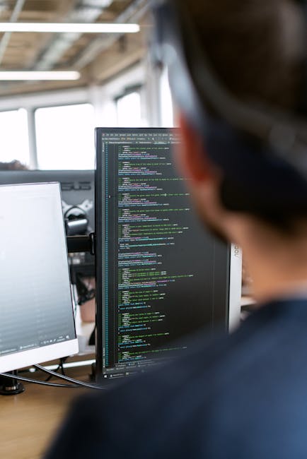 A close-up view of a person working outdoors with a computer, featuring a large monitor displaying lines of code. The background shows a garden with well-maintained grass, flower beds edged with neat 
