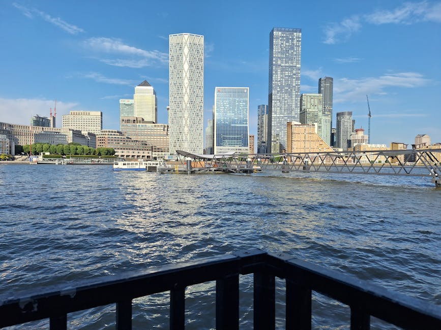 View of a modern city skyline across a body of water, with various tall skyscrapers featuring glass facades reflecting sunlight. In the foreground, a black metal railing borders a balcony or waterfron