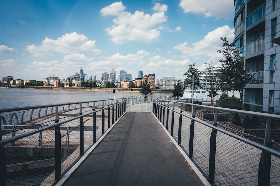 View of a modern city skyline across a body of water, with various tall skyscrapers featuring glass facades reflecting sunlight. In the foreground, a black metal railing borders a balcony or waterfron