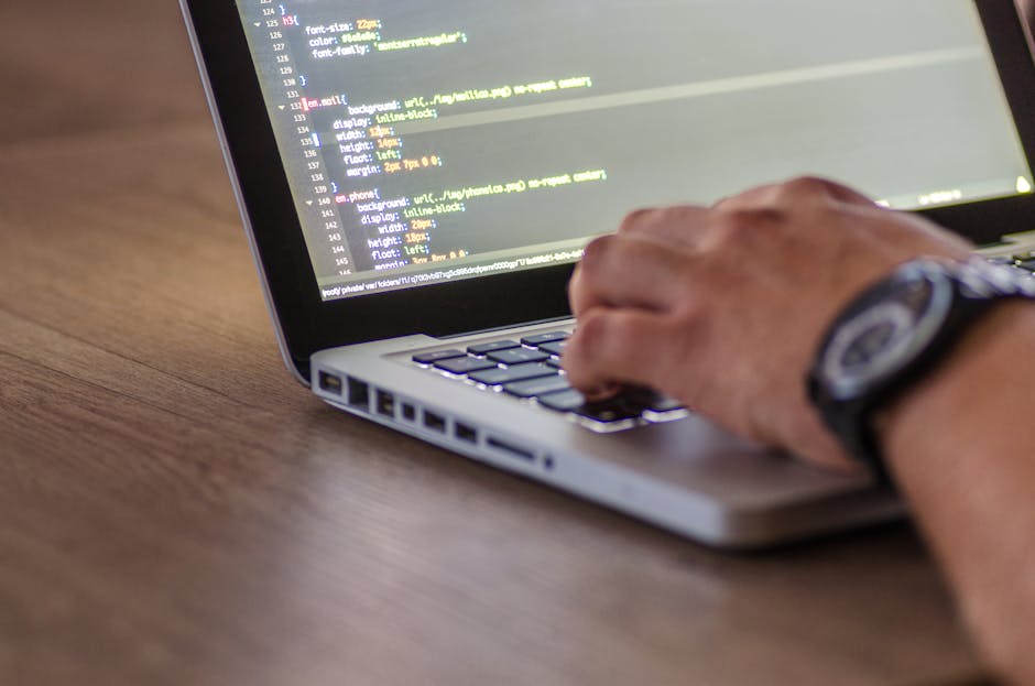 A close-up view of a person's hand working on a laptop placed on a wooden surface, with the screen displaying lines of code in a text editor. The individual is wearing a wristwatch, and the focus is o